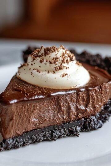 A slice of rich Mississippi mud pie with an Oreo cookie crust, topped with a swirl of whipped cream and a sprinkle of chocolate shavings, served on a white plate.
