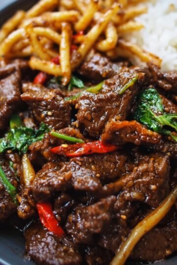 A cookedblack pepper beef served with rice and noodles, featuring dark, caramelized beef pieces, red chili, basil, and sesame seeds.